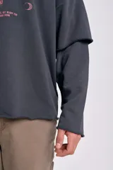 Sudadera de manga larga color gris oscuro con efecto desgastado y dobladillo sin rematar en el bajo. Presenta un diseño de doble manga, donde la manga interior es visible en el puño. Estampado frontal en color rosado con texto temático.