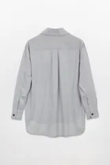 Camisa gris claro de tela Oxford, corte amplio, con cuello clásico, cierre frontal con botones negros, manga larga con puños abotonados, bolsillo de parche en el pecho y ruedo redondeado.