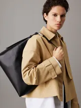 Cartera tipo hobo de piel sintética negra, con silueta fluida y correa ajustable con herrajes metálicos. Presenta cierre de cremallera principal y logo Calvin Klein grabado en la parte delantera.
