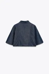Capa corta de denim azul con cuello alto y mangas amplias. Cierre frontal con cremallera metálica plateada.
