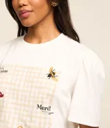 Remera blanca de corte slim con cuello redondo y mangas cortas abullonadas. Presenta un estampado frontal con diseño de picnic en tonos beige, con detalles bordados de abeja, cesta, hongo y texto en francés.