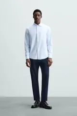 Camisa celeste a rayas verticales blancas, de corte recto, con cuello italiano abierto, mangas largas con puños abotonados y cierre frontal con botones.