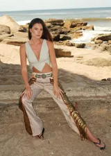 Pantalón de tiro medio con corte acampanado y tajos laterales. El tejido principal es brocado en tonos claros con un patrón sutil, y presenta apliques de lentejuelas doradas a lo largo de los tajos laterales. Incluye un cinturón ancho de cuero claro con hebilla metálica de estilo western.