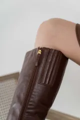 Bota de cuero color marrón con caña alta, taco cuadrado y punta fina. Presenta una correa con ojales metálicos dorados alrededor del tobillo.