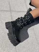 Bota negra de ecocuero con caña media, plataforma track y tres correas con hebillas plateadas.