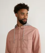 Camisa overshirt color salmón de sarga de algodón con capucha gris ajustable con cordones. Tiene cierre frontal con botones, dos bolsillos con solapa en el pecho y mangas largas con puños abotonados.