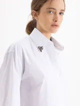 Camisa blanca de poplin de algodón con fit cómodo y largo que cubre hasta debajo de la cintura. Presenta botones ocultos en cartera y puños, y punteras de níquel en el cuello.