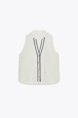 Camisa blanca con lunares negros, cuello redondo y escote pico. Sin mangas, con detalle de ribete negro en el escote y cierre frontal con botones.