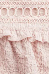 Top sin mangas color rosa pálido con textura arrugada, escote en V con tiras para atar y detalles de encaje calado en el canesú. Presenta mangas cortas con volados y corte holgado con costura fruncida bajo el busto.