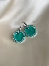 Aros colgantes con forma circular, hechos de resina color lila y borde de strass plateado. El aro es de acero quirúrgico plateado.