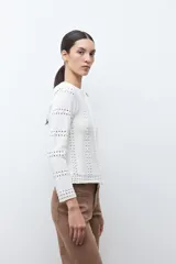 Chaqueta de tejido punto color blanco crudo, de manga larga y cuello redondo, con cierre frontal metálico. Presenta un diseño calado con aberturas verticales a lo largo del cuerpo y las mangas.