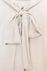 Blusa color crudo de tejido satinado, con cuello subido, manga sisa y cierre en la espalda con lazada y botones.