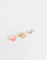 Set de tres anillos dorados de metal con diferentes diseños: uno con un corazón rosa, otro con forma de rosa y otro con una perla sintética.
