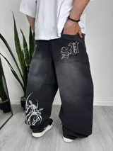 Pantalón baggy de jean negro con efecto lavado, con estampado de un esqueleto blanco en la pierna izquierda y bordado de letras góticas en el bolsillo trasero derecho.