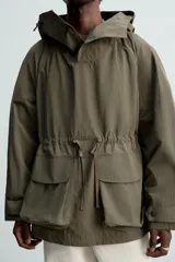 Parka color verde militar de corte amplio, confeccionada en tejido técnico ligero. Cuenta con cuello alto con capucha ajustable, cierre frontal con botones, mangas largas con puños ajustables, bolsillos de plastrón con solapa en la parte delantera, cintura ajustable con cordón y bajo con aberturas laterales ajustables con cordón.