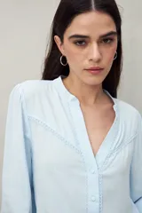 Camisa celeste de manga larga con cuello redondo y escote en V. Presenta detalles de pasamanería en el frente y las mangas, y botones forrados al tono. De corte fluido.
