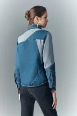 Campera recta de denim rígido con combinación de recortes en diferentes tonos de azul, cuello camisero, cierre frontal con botones metálicos, bolsillos con solapa y botones metálicos en el pecho, y puños con detalle de encaje negro.