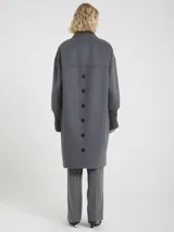 Abrigo largo de color gris confeccionado a mano, con cuello y puños acanalados. Presenta un diseño de corte recto con cierre frontal de botones, bolsillos laterales y botones decorativos en la parte posterior.