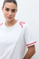 Remera blanca de algodón con cuello redondo y mangas cortas. Presenta bordados florales rojos en los hombros y en los bordes de las mangas.