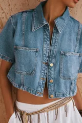Camisa de denim celeste de corte crop, con cuello solapa, escote en pico, manga corta y dos bolsillos delanteros con botón. Cierre frontal con botones metálicos.