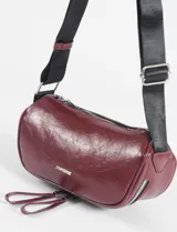 Riñonera cilíndrica de cuero vegano color borgoña, con dos compartimentos separados con cierres plateados. Posee una correa ajustable de tela negra y tiradores de cuero en las cremalleras.