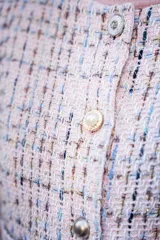Chaqueta corta tipo Chanel de tweed color rosa con detalles en blanco y azul. Tiene cuello redondo, cierre frontal con botones perlados y bolsillos delanteros.
