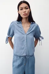 Camisa celeste de denim con cuello solapa, escote en pico, mangas abullonadas con lazos y cierre frontal con botones.