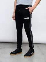 Pantalón deportivo Umbro UNY de piqué negro con puños ajustados, cintura elástica con cordón y logo Umbro estampado en blanco. Presenta una franja gris con ribete blanco a lo largo de la pierna. Confeccionado en poliéster y algodón.