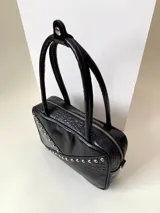 Cartera rígida negra de cuero con tachas plateadas y tres combinaciones de cuero diferentes que aportan textura. Tiene herrajes y cierre metálico plateado marca YKK. Cuenta con un asa fina para llevar al hombro o en la mano y una correa larga extra regulable.