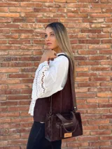 Cartera bandolera de cuero color marrón chocolate, con forro de tela y herrajes dorados. Tiene doble asa de mano y correa larga regulable y desmontable. Incluye bolsillo frontal con cierre y bolsillo interno.