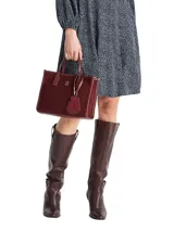 Cartera tipo tote bag de color borgoña, con acabado liso y detalles en cuero sintético del mismo tono. Presenta dos asas superiores y una correa ajustable extraíble. Incluye un colgante decorativo con el escudo de Hilfiger bordado y un dije metálico con el monograma TH.