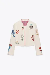 Chaqueta corta color crema con cuello redondo y mangas largas. Presenta bordados de motivos florales, corazones, cerezas, tazas y una silla. Tiene forro interior a rayas verticales rosas y blancas.