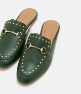 Zapatilla mule color verde oscuro, con puntera redonda, bridón metálico dorado, tachas doradas y suela marrón.