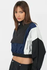 Buzo rompevientos anorak gris, de cuello alto, silueta boxy fit, con elástico en terminación de cintura y mangas. Detalle de recortes a contratono. Cierre centrado en delantero de acceso. Bolsillo con tapa por debajo, con cierre escondido. Tancas en cuello con elástico en su contorno, por interior. Estampa en lateral derecho, y centrado en espalda.