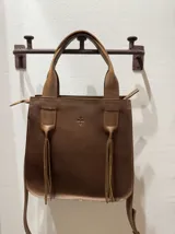 Cartera de cuero vacuno color marrón, con doble asa corta y correa larga desmontable. Tiene cierre superior y forro interior. Presenta detalle de flecos en los laterales y logo grabado en el centro.