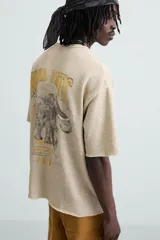 Camiseta color beige de punto tejido en hilatura de algodón, corte relaxed fit, cuello redondo y manga corta. Estampación con efecto lavado combinada a contraste en espalda. Acabados irregulares.