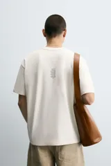 Camiseta color crudo de corte holgado, cuello redondo y manga corta. Tiene estampado de salpicaduras de pintura negra en el frente y texto estampado en la espalda.