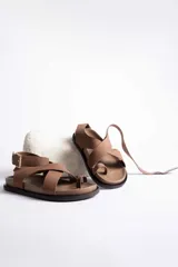 Sandalia plana de cuero color marrón topo, con tiras cruzadas en el empeine y tira sujetadora con hebilla dorada alrededor del tobillo. Posee una suela gruesa de goma negra.