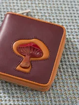 Billetera pequeña de cuero vacuno color bordó con cierre metálico y aplique de hongo. Interior con divisiones para tarjetas y forrería de tejido jacquard.