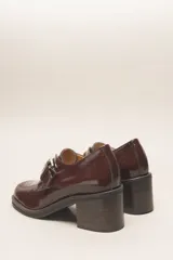 Zapato de cuero vacuno charol negro, con punta redondeada y detalle de perforado en la capellada. Tiene una tira con hebilla metálica y un taco cuadrado de 4 cm.