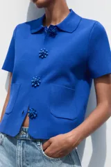 Chaqueta de punto azul con cuello solapa y manga corta. Tiene bolsillos delanteros de parche y cierre frontal con botones grandes con abalorios.
