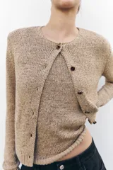 Chaqueta color beige de punto con cuello redondo, manga larga, rib en los bordes y cierre frontal con botones negros.