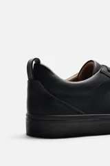 Zapatilla urbana de cuero sintético color negro, con cordones y suela plana.