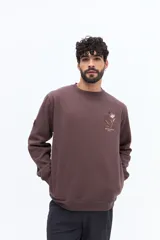 Buzo de algodón color marrón oscuro (ciruela/ciruela quemado) con cuello redondo, puños y ruedo acanalados. Presenta un pequeño bordado de motivos florales en el pecho.