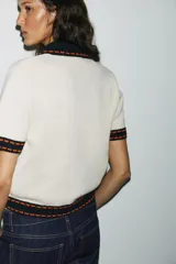 Chaqueta color blanco de punto con cuello solapa y manga corta. Tiene cierre frontal con botones color naranja y ribete en azul marino y naranja.