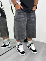 Bermuda de jean gris oscuro con efecto lavado y tachas plateadas distribuidas en la parte delantera de las piernas. Corte ancho y largo, estilo capri o culotte.