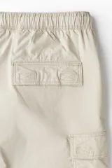 Short color beige de tejido grueso de algodón, con corte estándar, cintura elástica con cordón ajustable, bolsillos al bies y bolsillos de solapa con cierre autoadherente en las piernas y atrás.