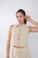 Top tejido color beige con espalda calada y cuello mao. Presenta cierre frontal con cordones y detalle de broches decorativos en el cuello.
