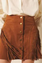 Minifalda de cuero gamuzado color marrón, con flecos y broches tipo snap en el centro delantero. Presenta tachas metálicas y remaches símil piedra a lo largo de la línea de los flecos.