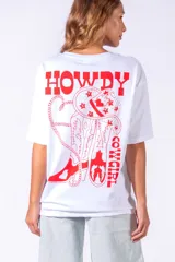 Remera blanca de corte holgado con estampado en la espalda que incluye un sombrero de cowboy, botas y la frase 'Howdy Cowgirl' en color rojo.